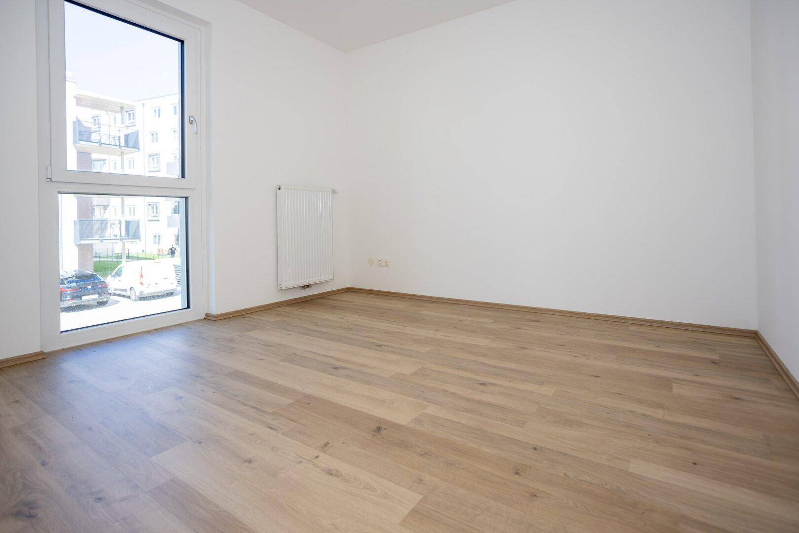Schlafzimmer mit hellem Laminatboden, weißen Wänden und großem Fenster mit Blick nach draußen.
