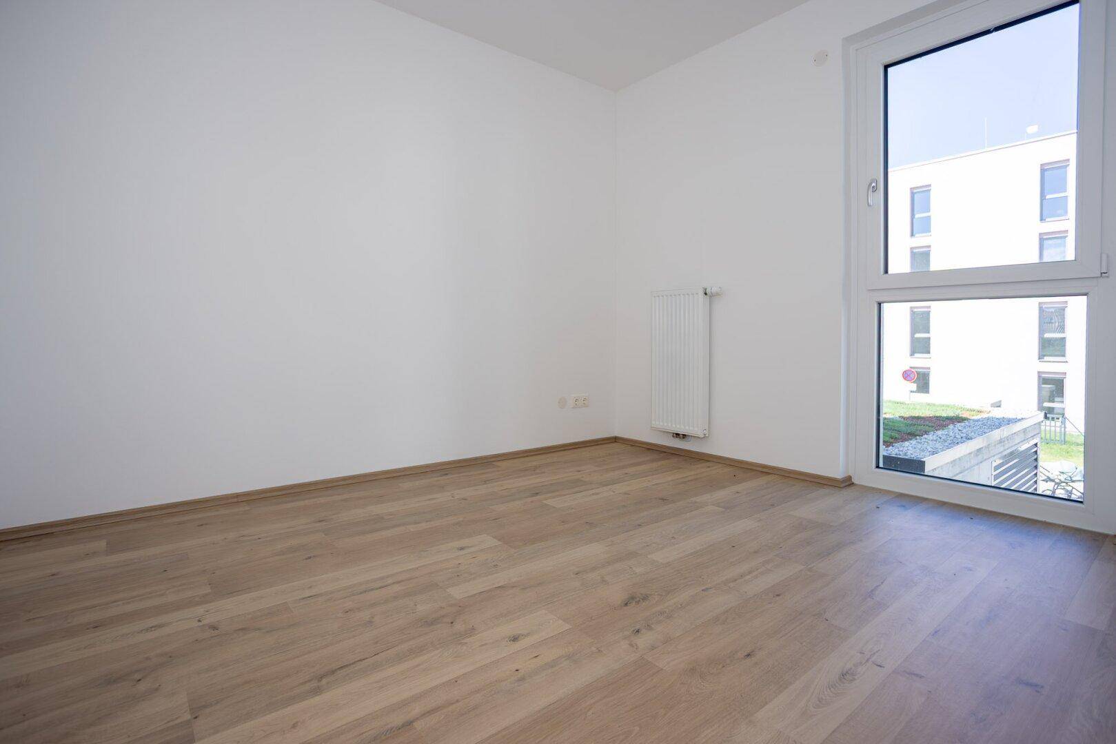Helles Schlafzimmer mit Laminatboden, weißen Wänden und Fenster mit Blick ins Grüne.