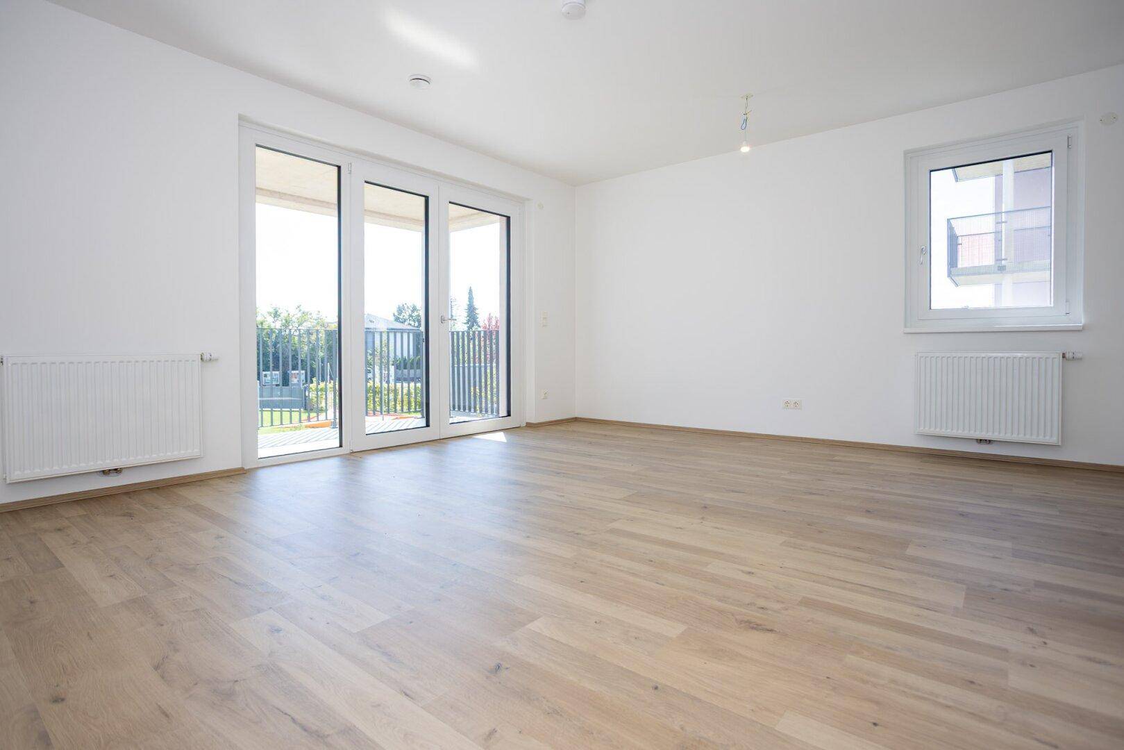 Helles Wohnzimmer mit modernem Laminatboden, weißen Wänden und Zugang zum Balkon.
