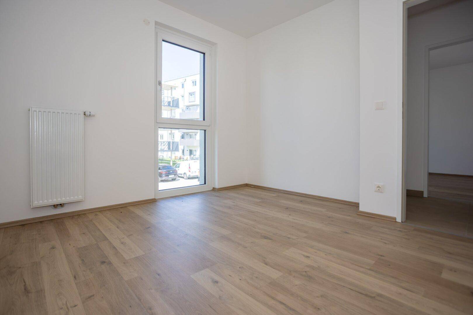 Schlafzimmer mit hellem Laminatboden, weißen Wänden und Fenster, das viel Tageslicht hereinlässt.