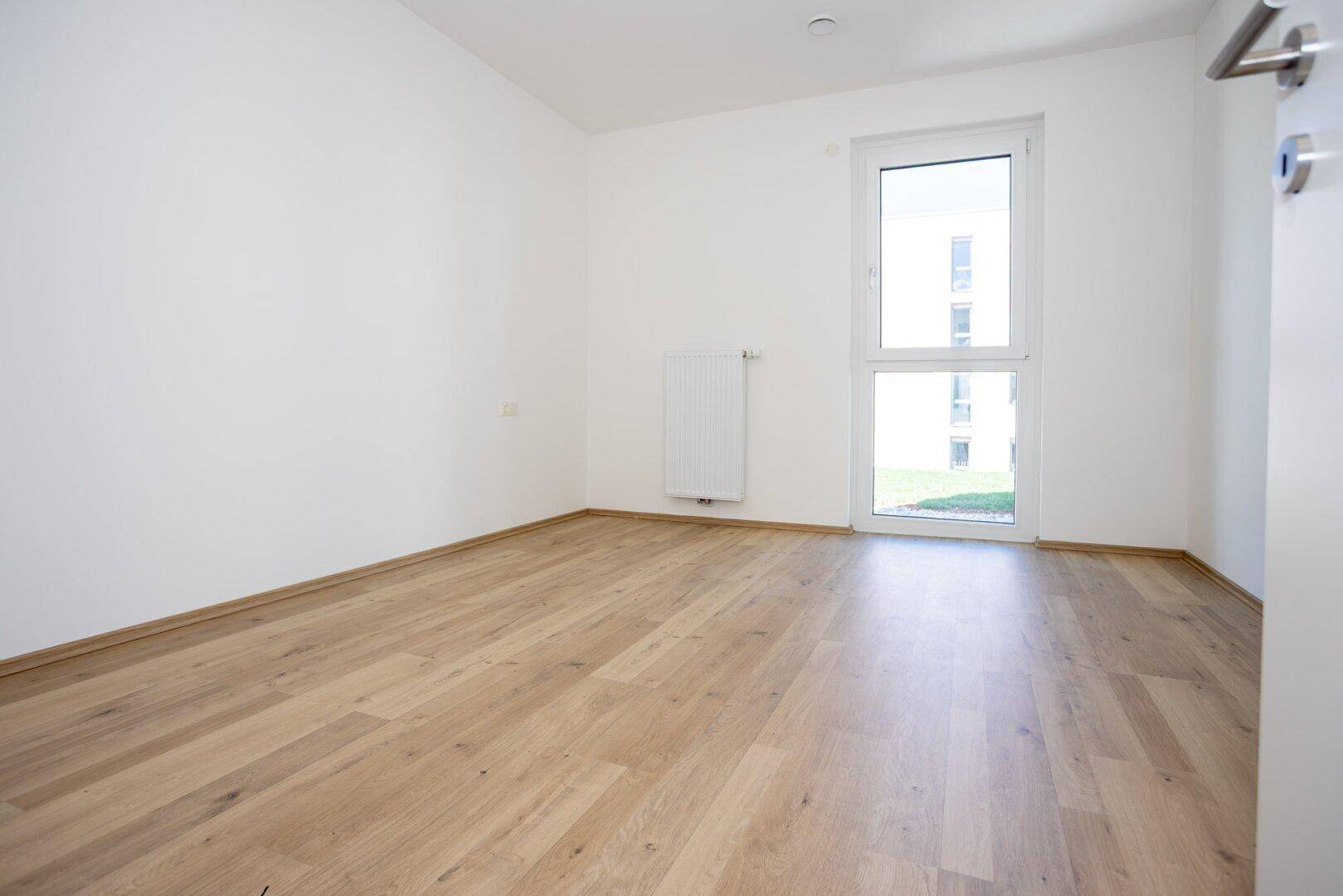 Helles Schlafzimmer mit Laminatboden, weißen Wänden und Fenster mit Blick auf die Außenanlage.