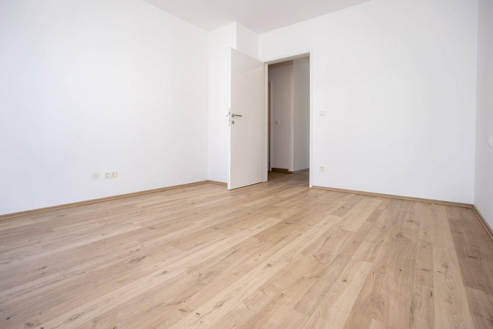 Leeres Schlafzimmer mit hellem Laminatboden und weiß gestrichenen Wänden, mit Zugang zu weiteren Räumen.