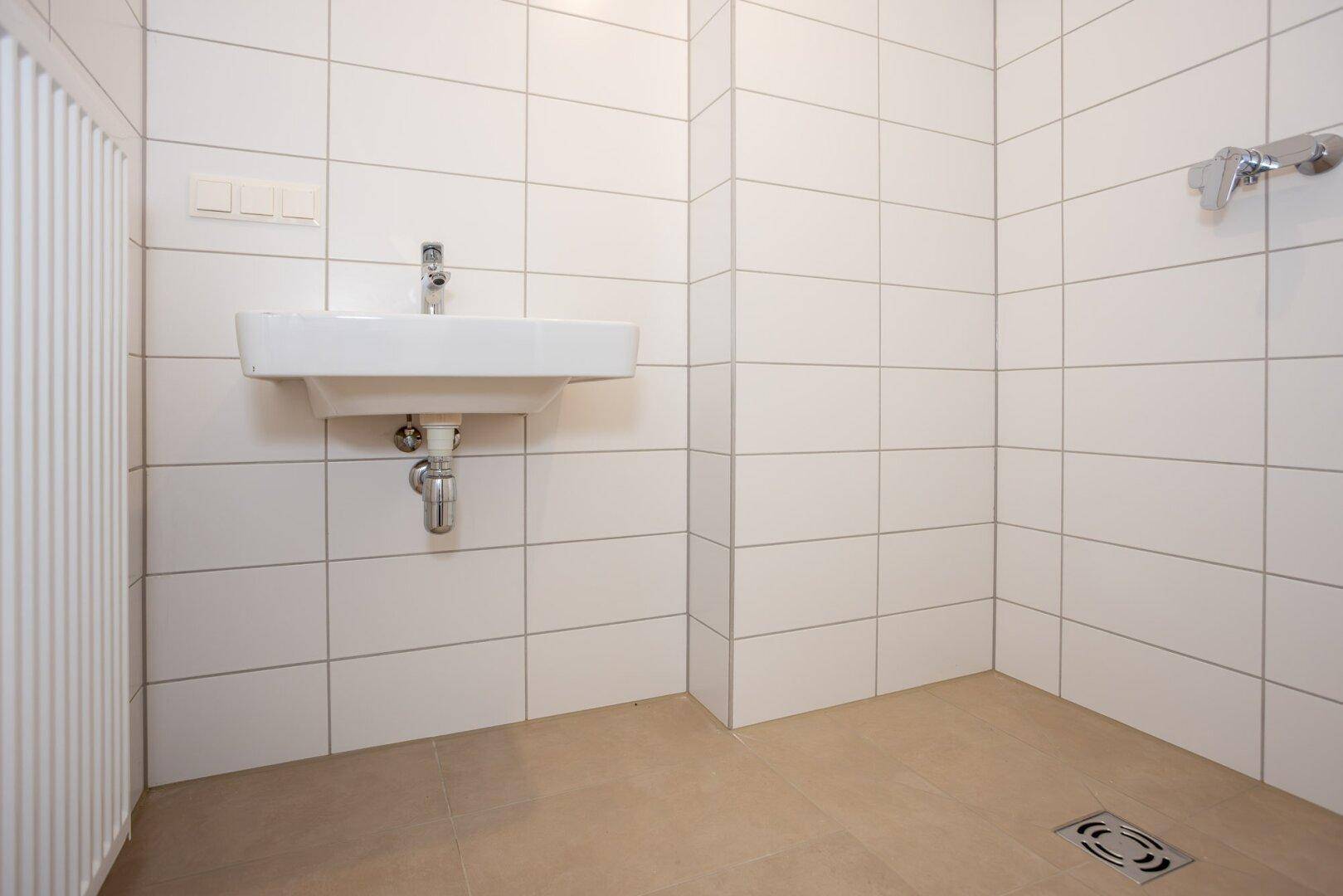 Modernes Badezimmer mit weißen Fliesen, beigefarbenem Boden, Waschbecken und Duschbereich.