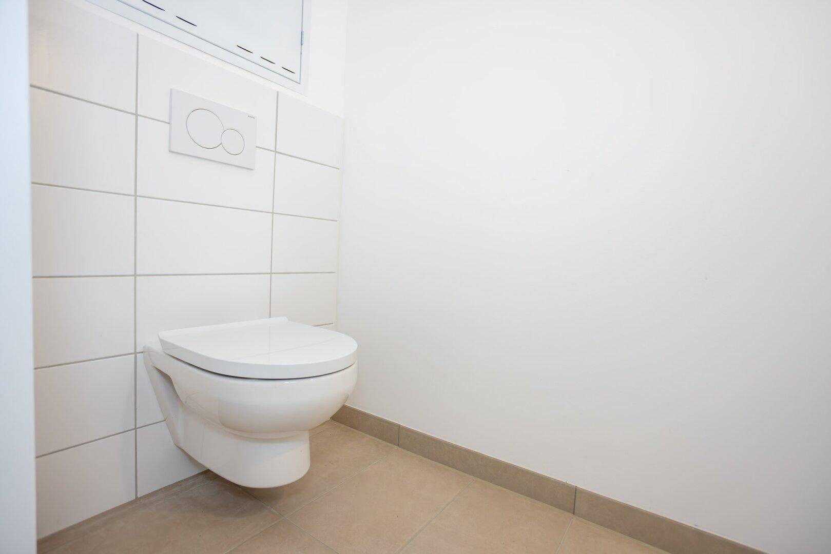 Separates WC mit wandhängender Toilette, weißen Wandfliesen und beigefarbenem Boden.