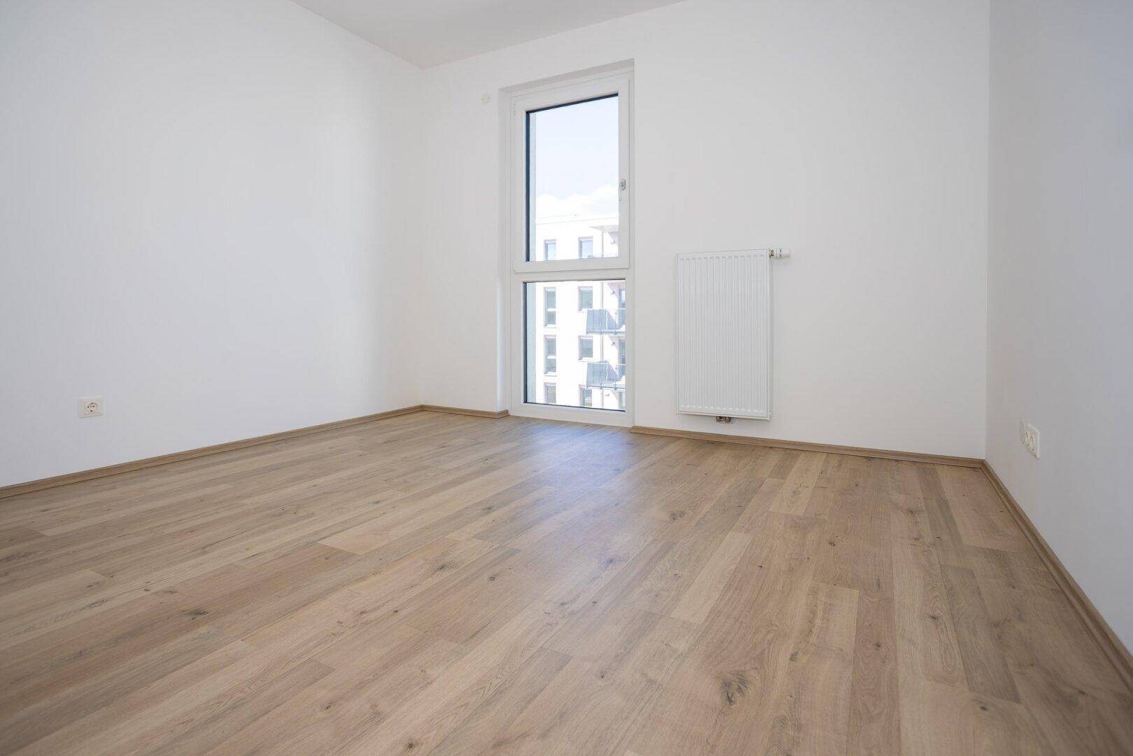 Helles Schlafzimmer mit Laminatboden und großem Fenster, das viel Licht hereinlässt.