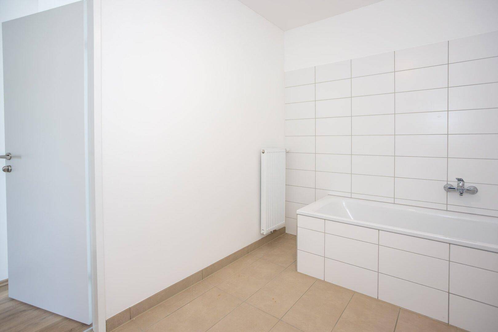 Helles Badezimmer mit weißen Wandfliesen, beigem Boden und einer Badewanne.