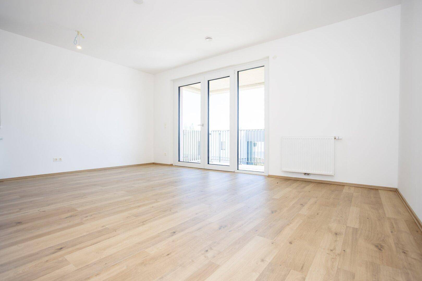 Helles, leeres Zimmer mit hellem Holzboden und Zugang zu einem französischen Balkon.