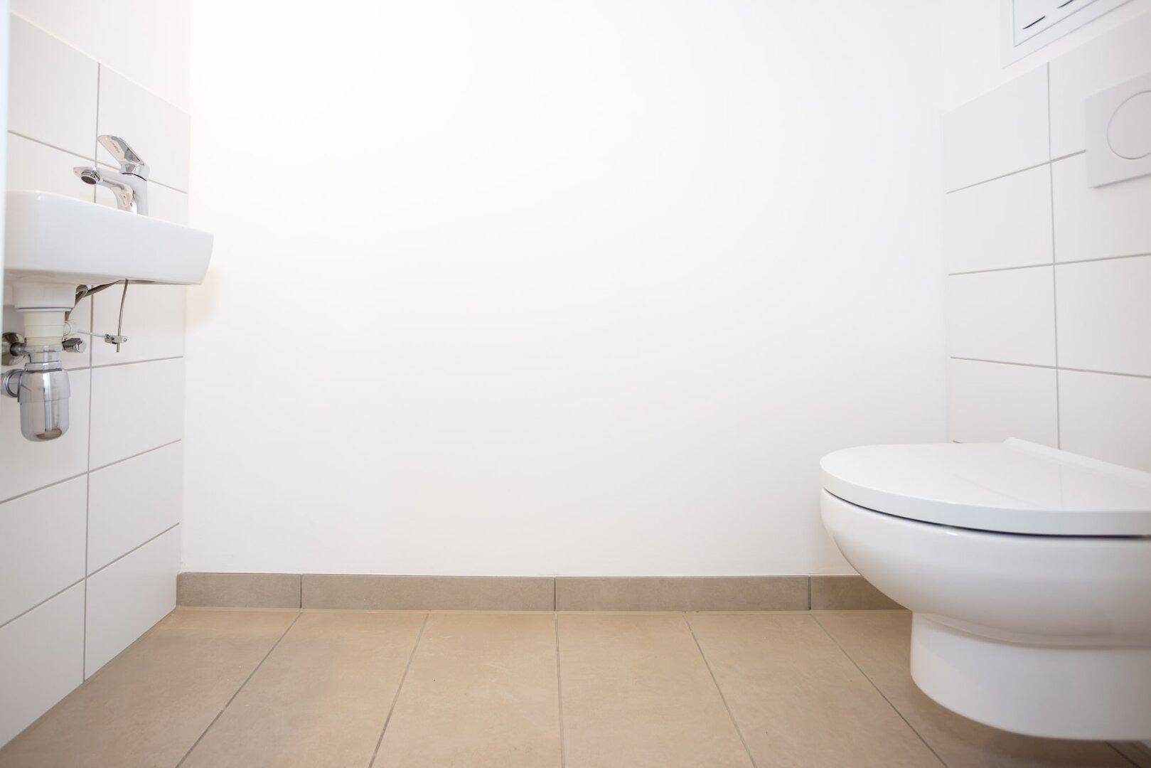 Separates WC mit weißen Fliesen, beigem Boden, Toilette und kleinem Waschbecken.