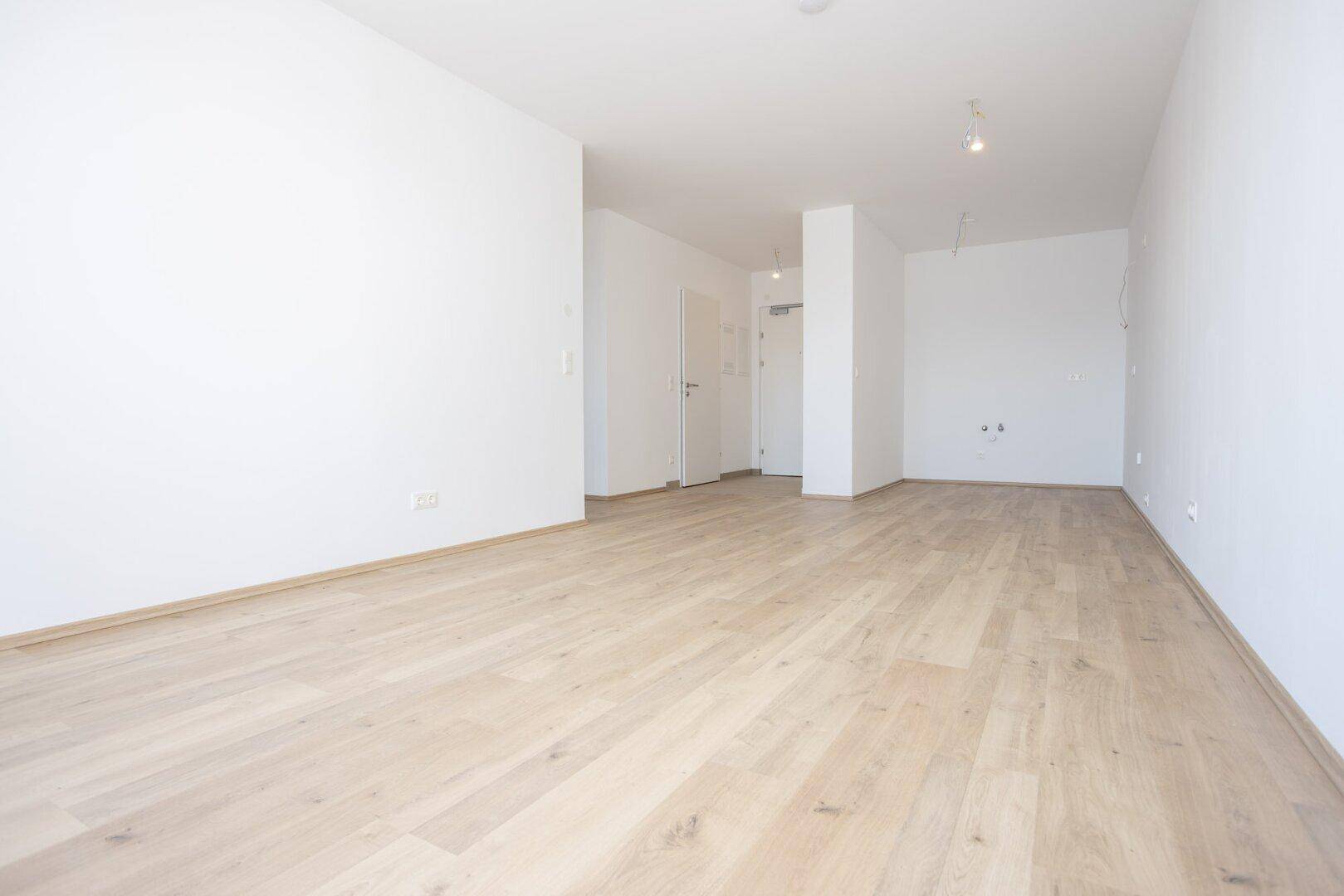 Geräumiger, unmöblierter Wohnbereich mit hellem Laminatboden und weiß gestrichenen Wänden.