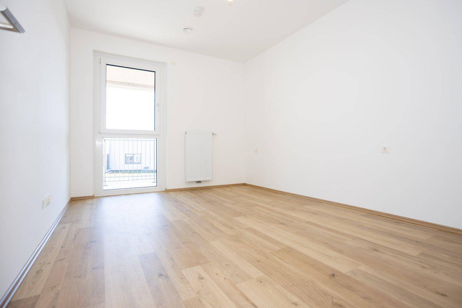 Helles, leeres Zimmer mit Laminatboden und großem Fenster, ideal als Schlaf- oder Arbeitszimmer.