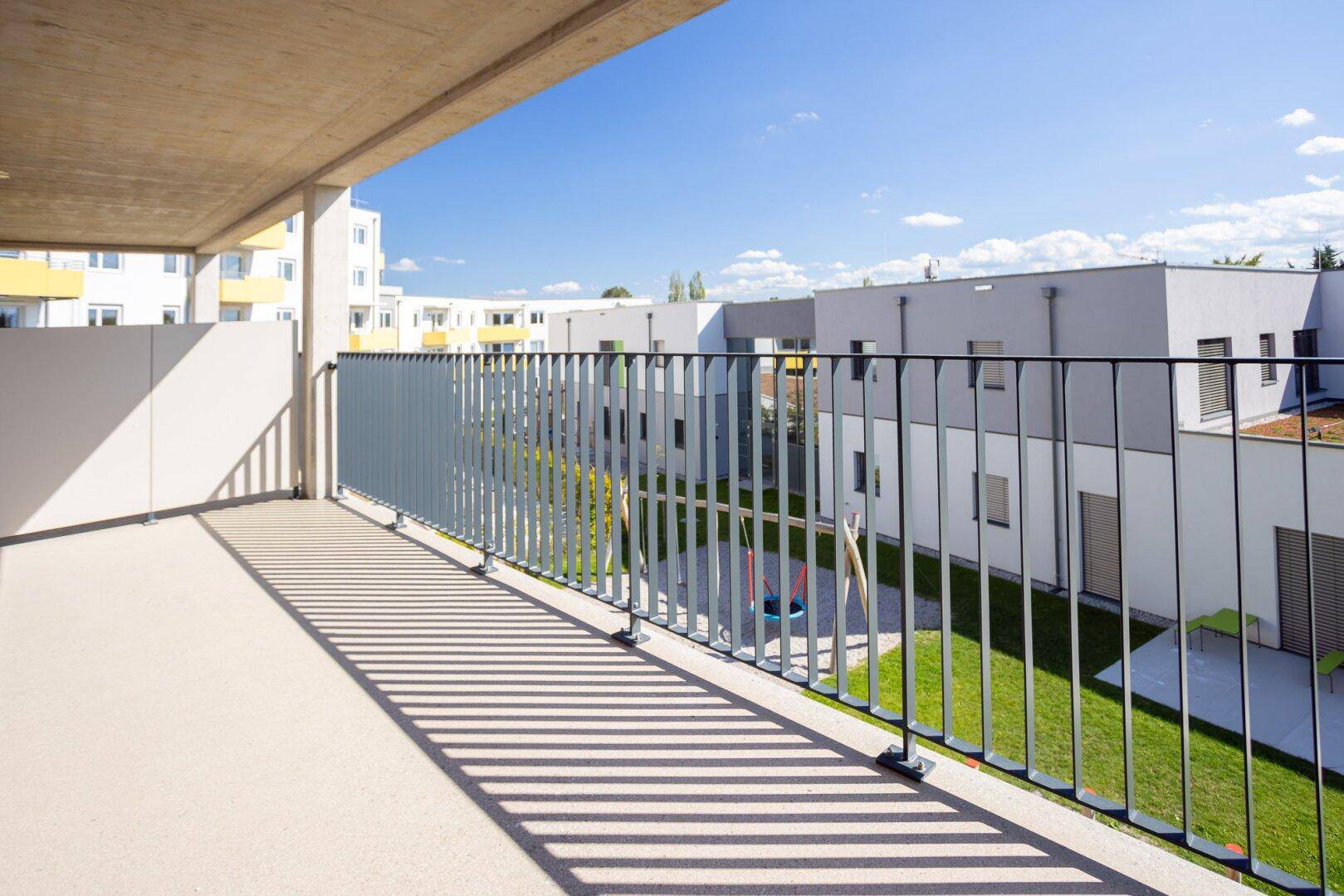 Sonniger Balkon mit Blick auf einen Innenhof mit Spielplatz und moderne Wohngebäude.