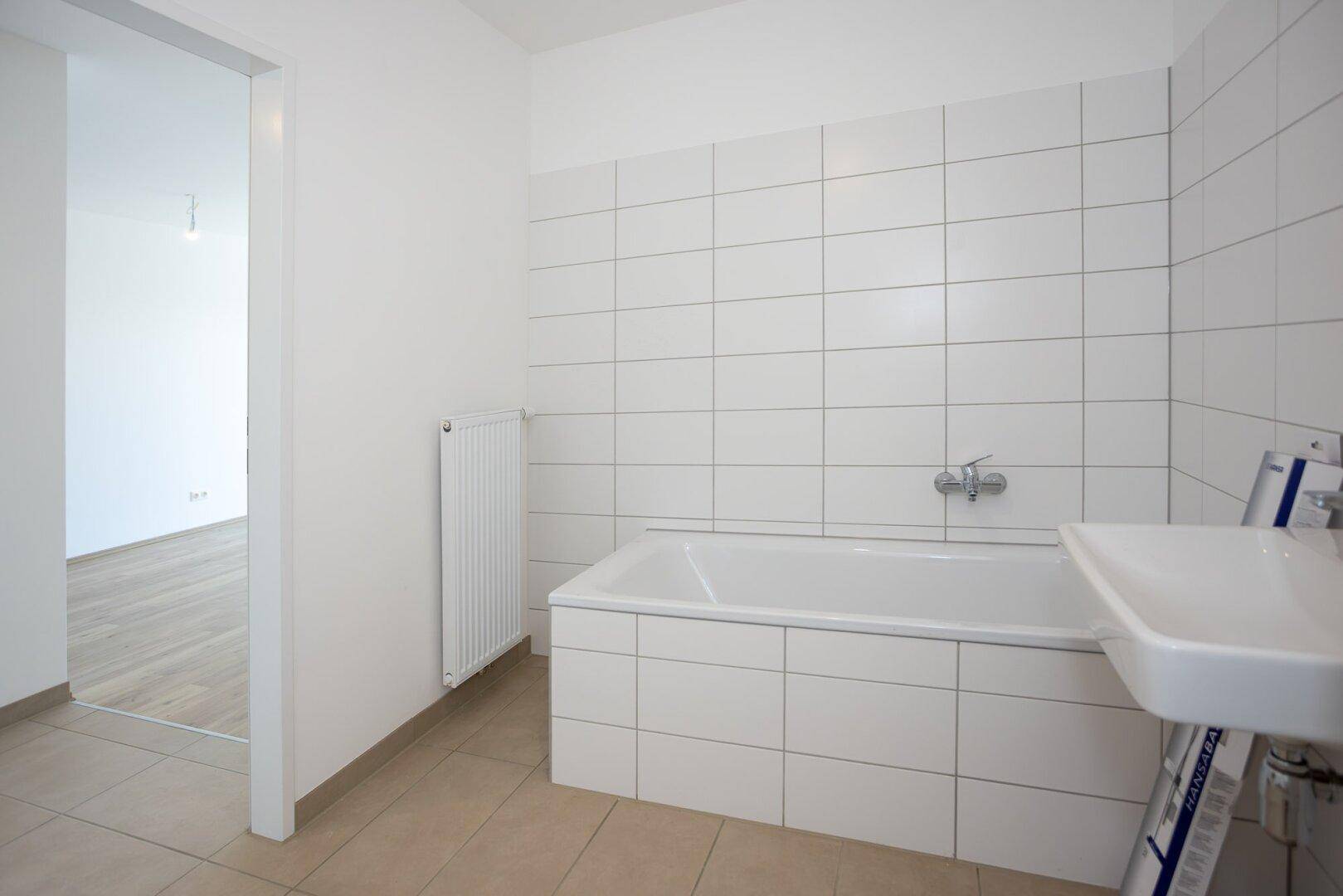 Helles Badezimmer mit Badewanne, Waschbecken und weißen Fliesen, mit Blick in den Wohnbereich.