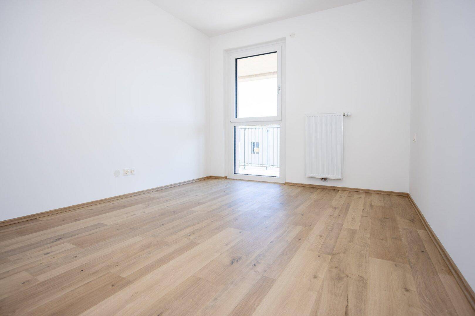 Unmöbliertes Zimmer mit hellem Laminatboden und weiß gestrichenen Wänden, mit Fenster.