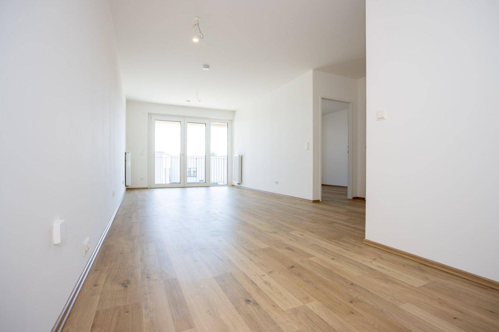 Heller, leerer Wohnraum mit hellem Laminatboden und Zugang zu einem französischen Balkon.