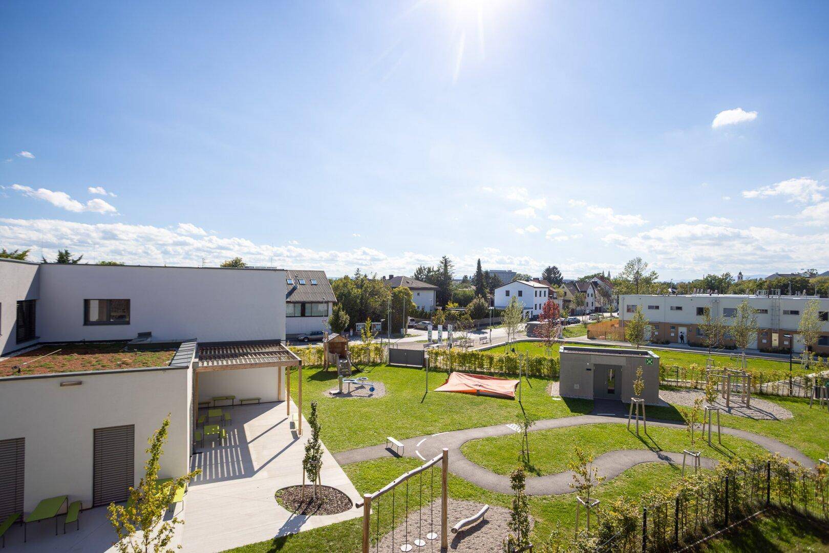 Weitläufige Außenanlage eines Neubaus mit Spielplatz und viel Grün unter strahlend blauem Himmel.