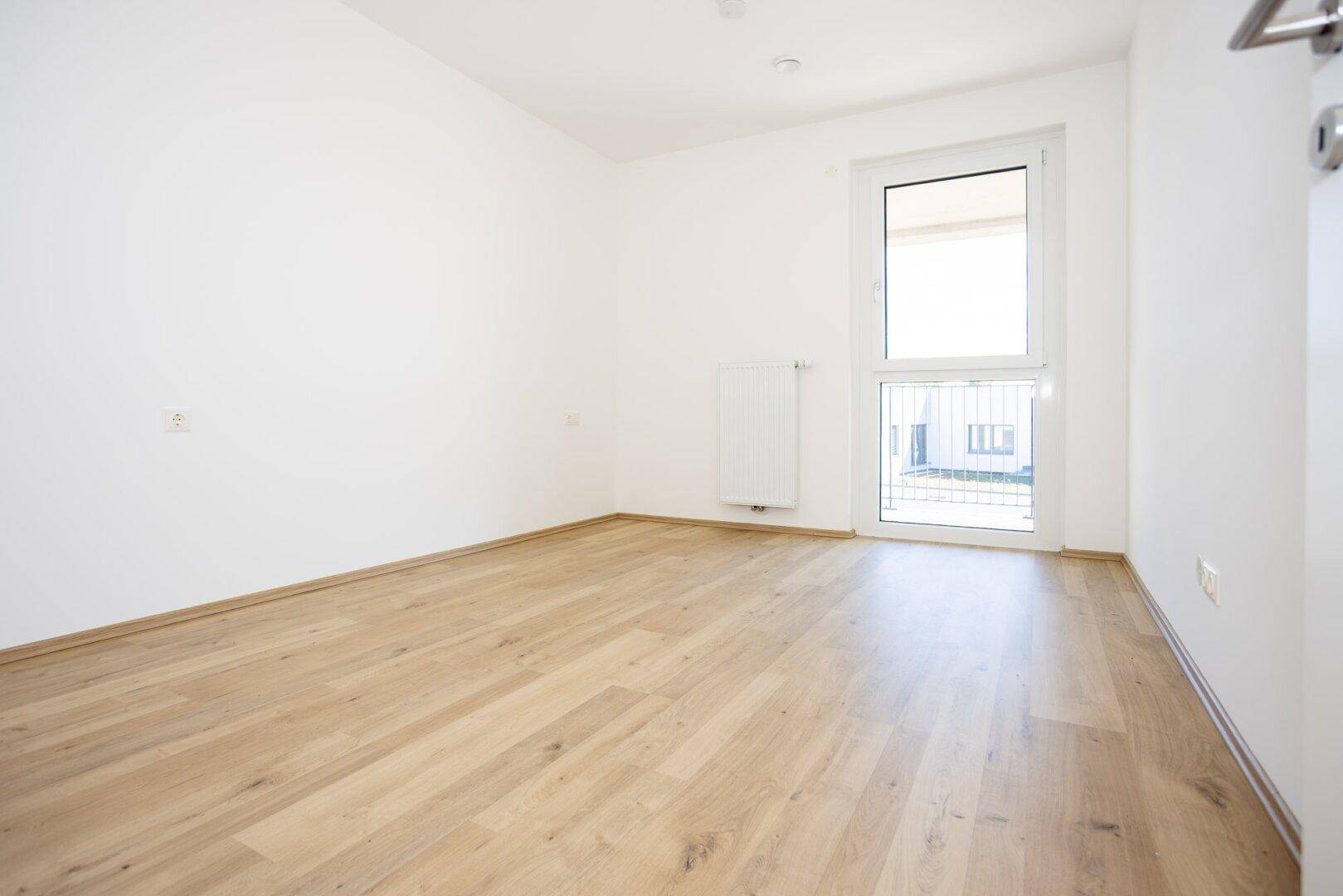 Helles Schlafzimmer mit Holzboden und großem Fenster, das viel Tageslicht hereinlässt.