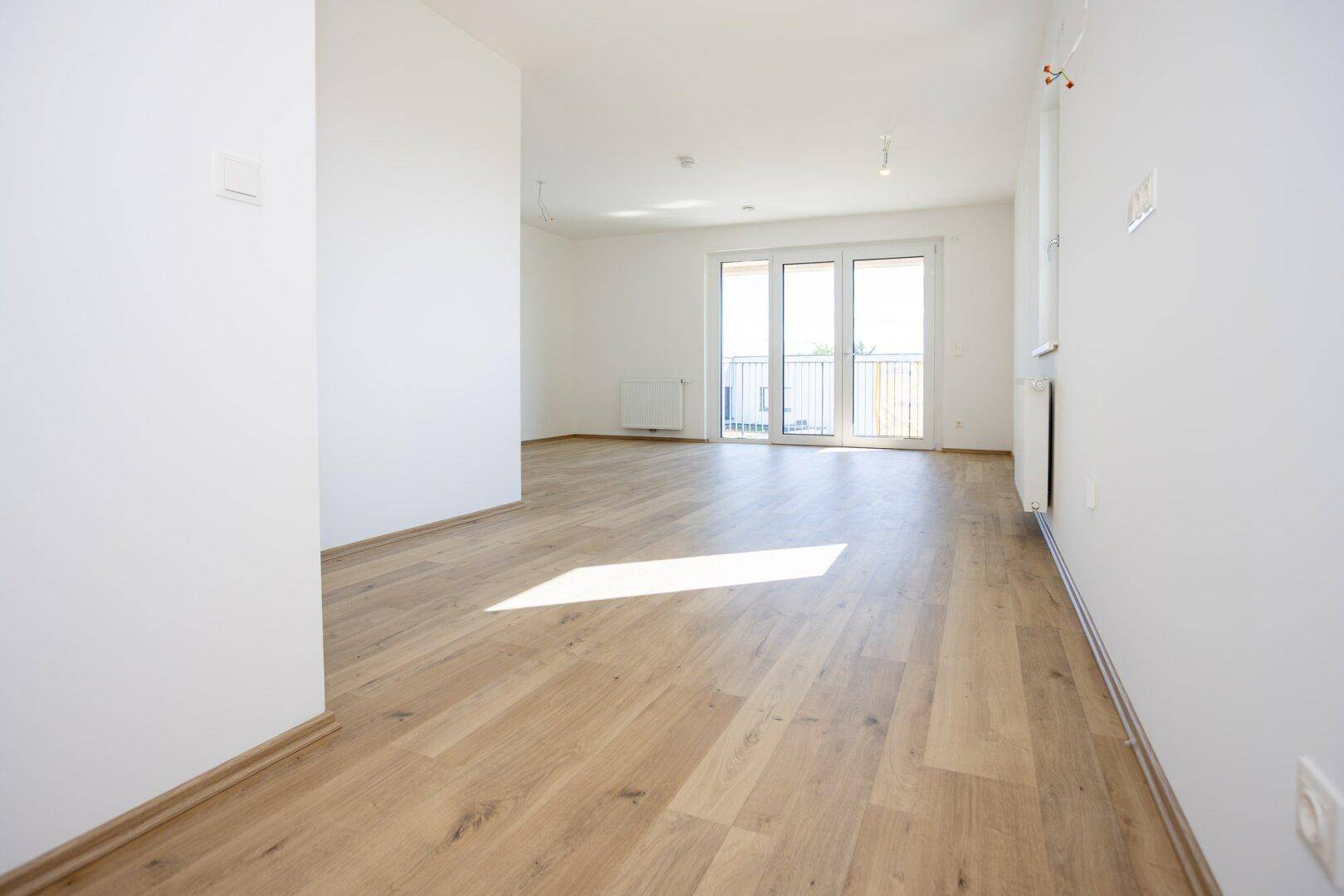 Lichtdurchfluteter Wohnbereich mit hellem Laminatboden und großen Fenstern zum Balkon.