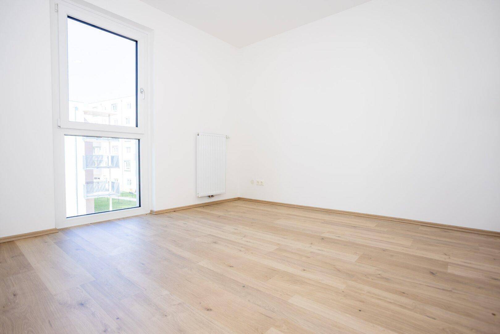 Helles Schlafzimmer mit Laminatboden und großem Fenster mit Blick ins Grüne.