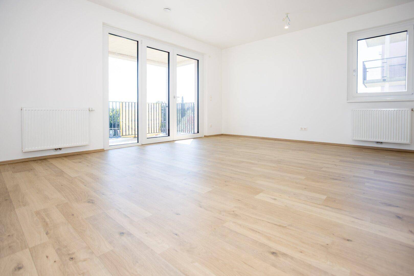 Geräumiges, helles Wohnzimmer mit hellem Laminatboden und Zugang zum Balkon.