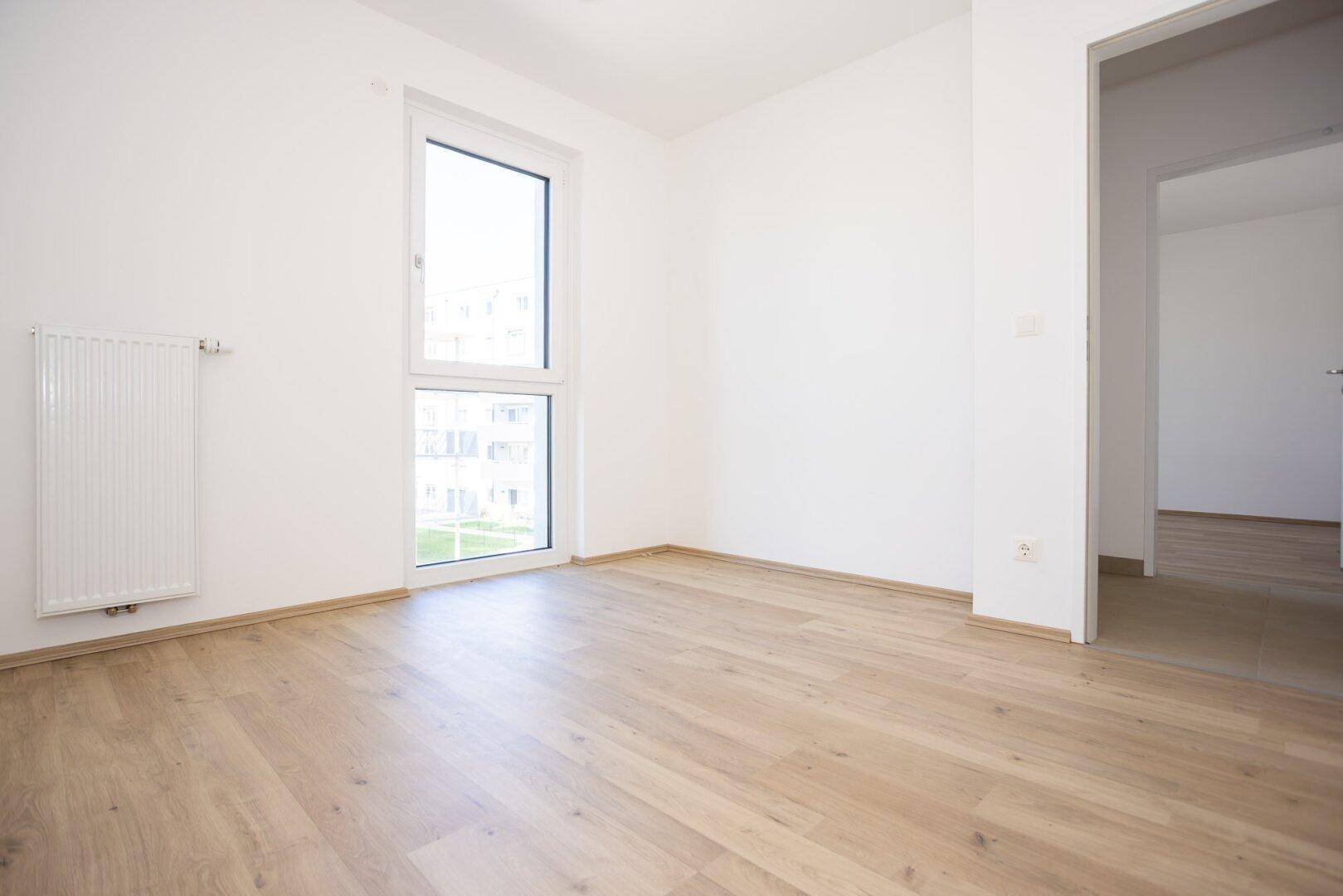 Helles Schlafzimmer mit Laminatboden und Blick auf die umliegenden Gebäude und Grünflächen.