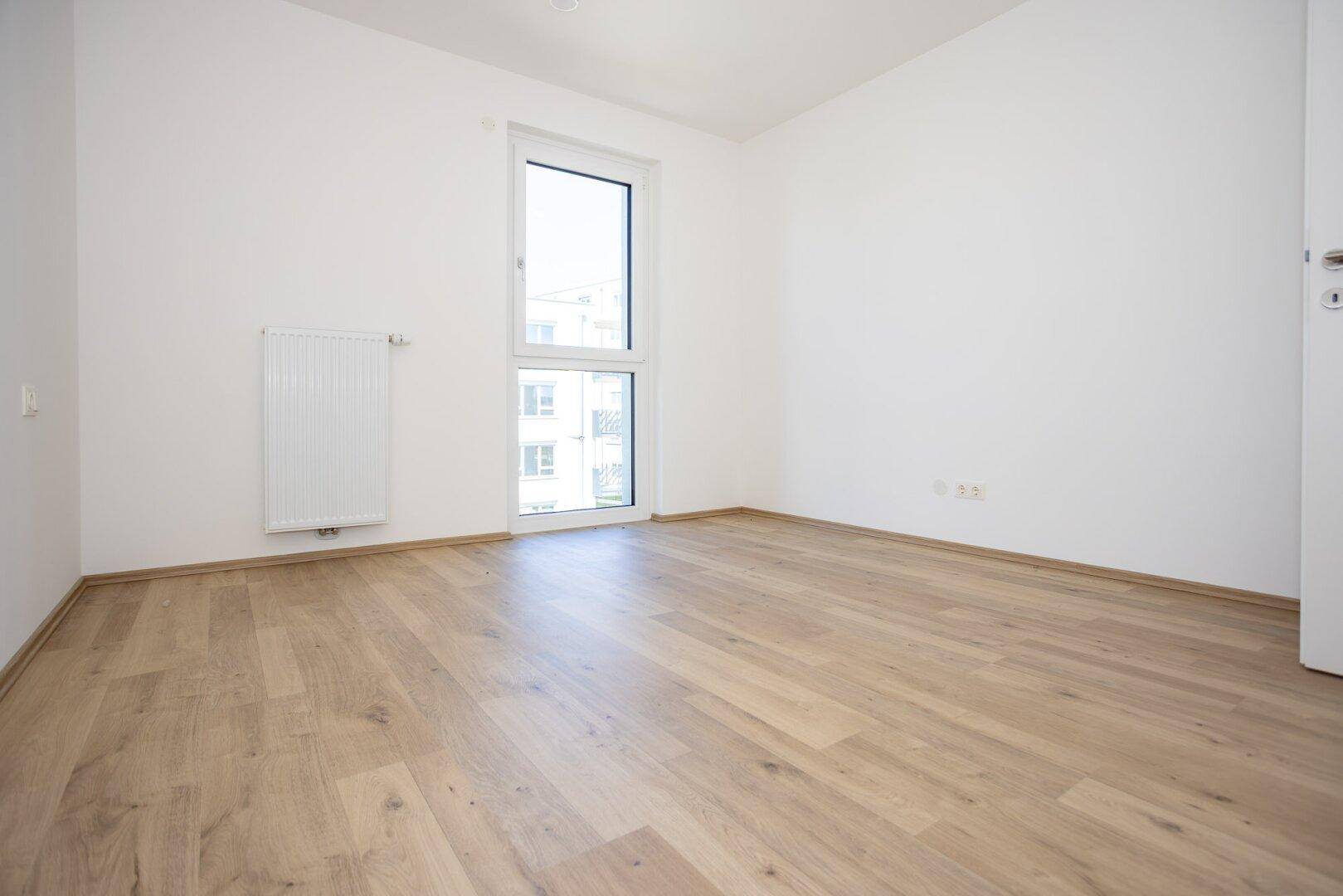 Helles Schlafzimmer mit Laminatboden und Blick auf die umliegenden Gebäude und Grünflächen.