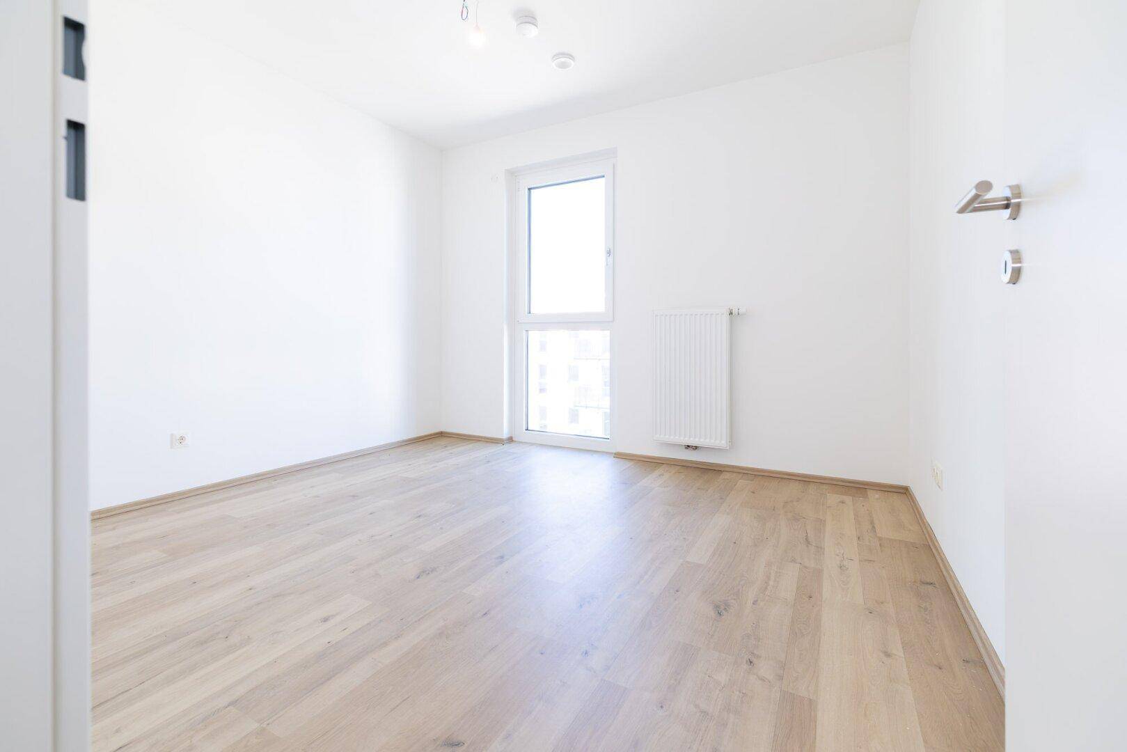Helles Schlafzimmer mit Laminatboden und großem Fenster, das viel Tageslicht hereinlässt.
