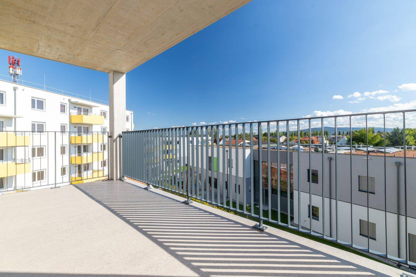 Breiter Balkon mit Metallgeländer und Ausblick auf die Wohnanlage unter blauem Himmel.
