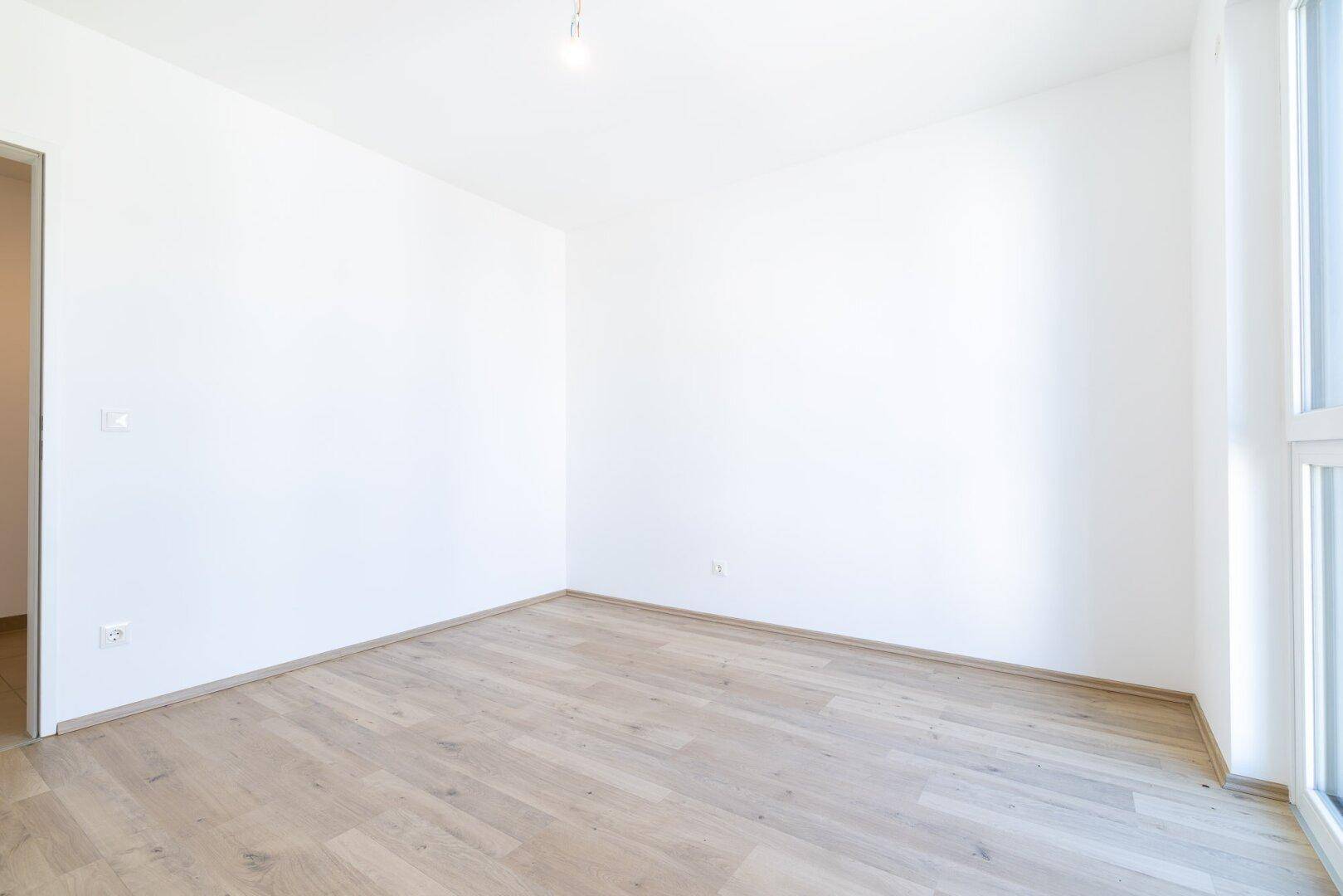 Leeres Zimmer mit hellem Laminatboden und weiß gestrichenen Wänden, ideal als Kinder- oder Arbeitszimmer.