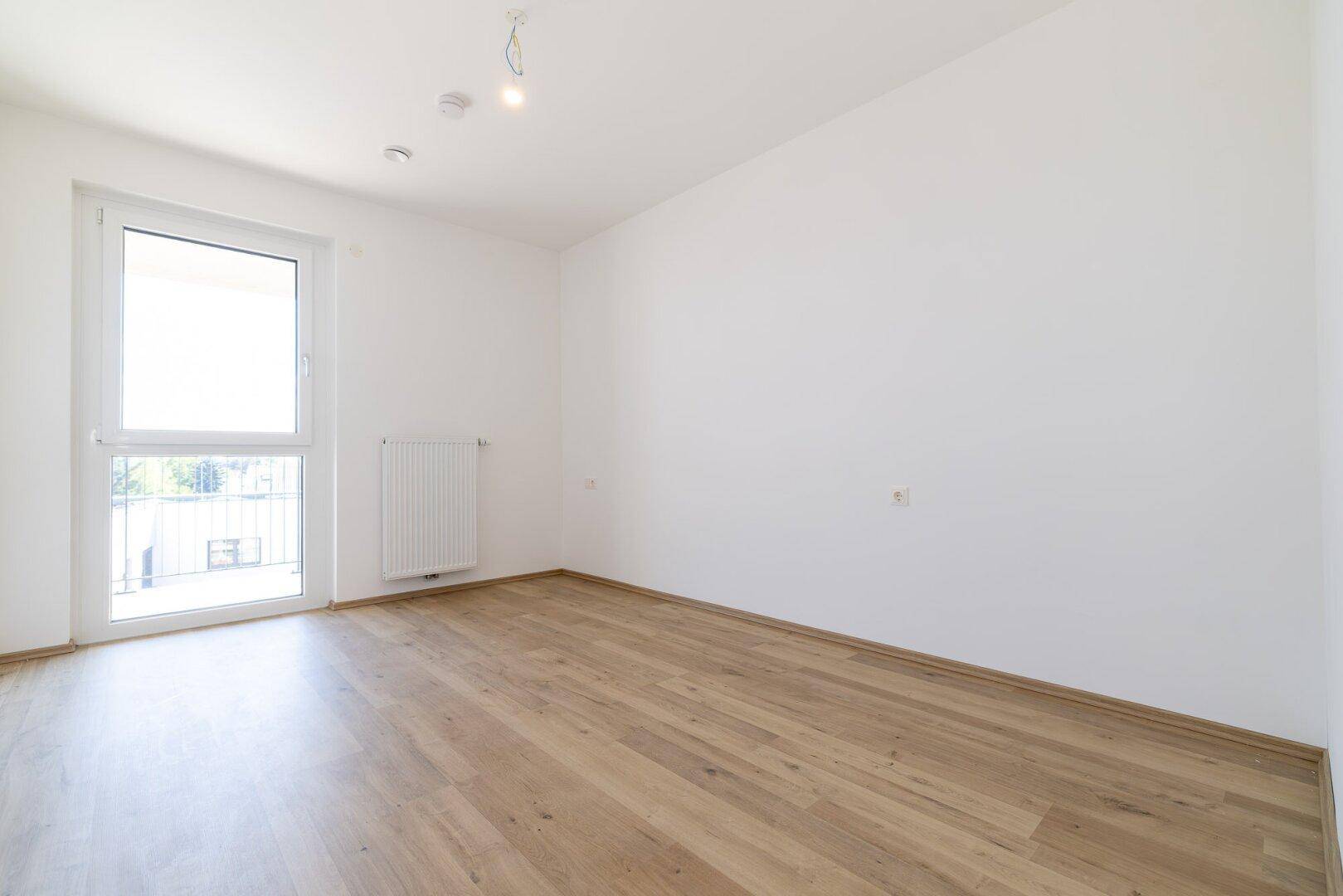 Helles Schlafzimmer mit Laminatboden und Fenster mit Zugang zu einem Balkon.