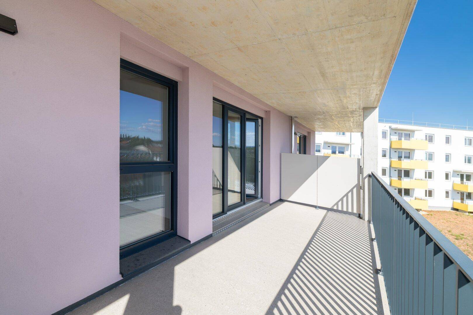 Langer Balkon mit hellrosa Fassade und Blick auf die Nachbargebäude unter blauem Himmel.