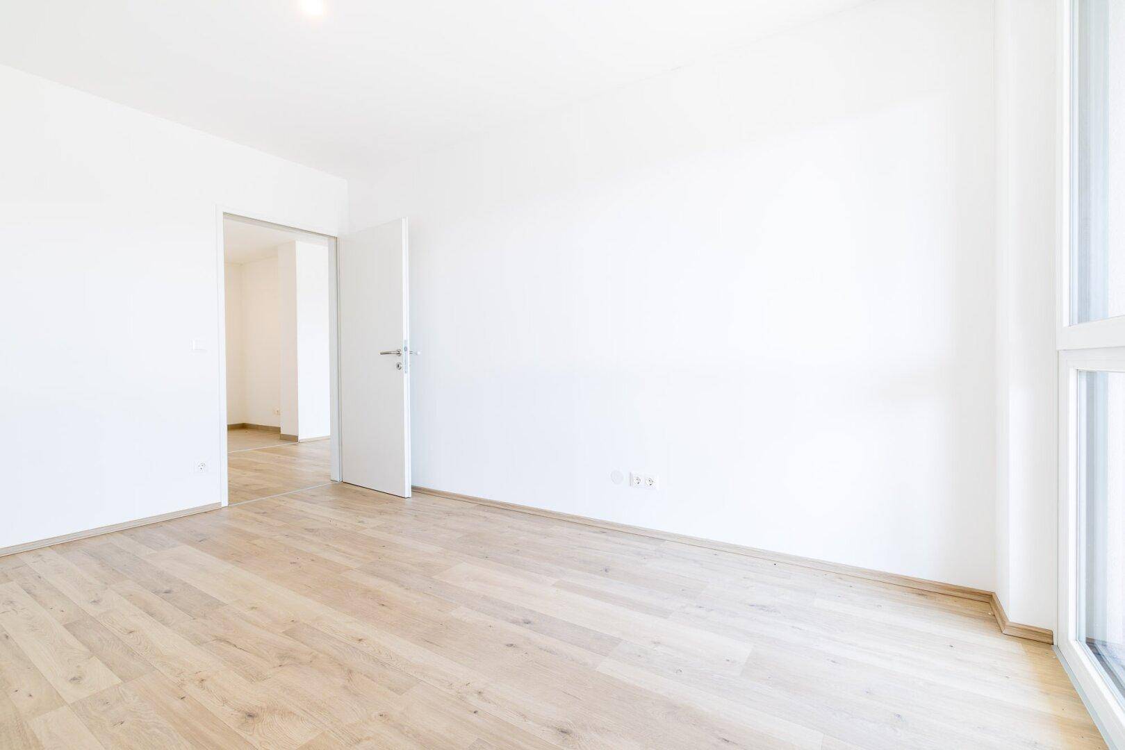 Heller, leerer Raum mit Laminatboden und offener Tür zu einem weiteren Zimmer.
