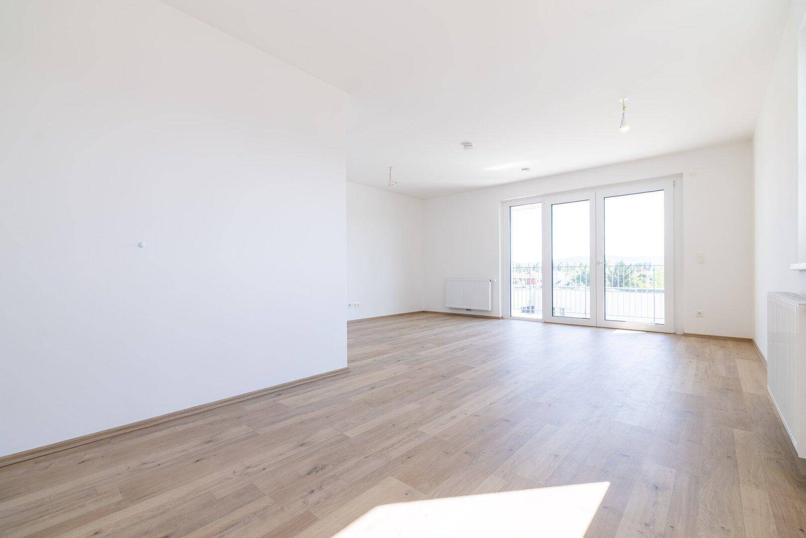 Helles, leeres Wohnzimmer mit hellem Laminatboden und Zugang zu einem Balkon.