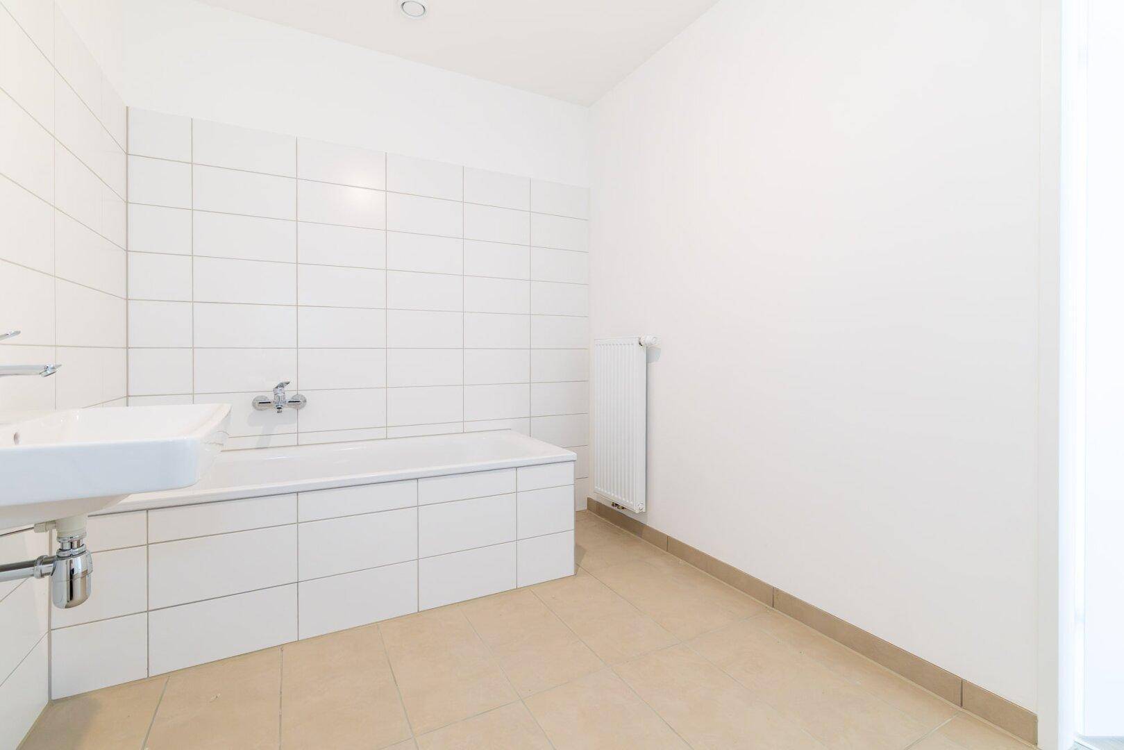 Helles Badezimmer mit weißen Wandfliesen, Badewanne und Waschbecken, unmöbliert.