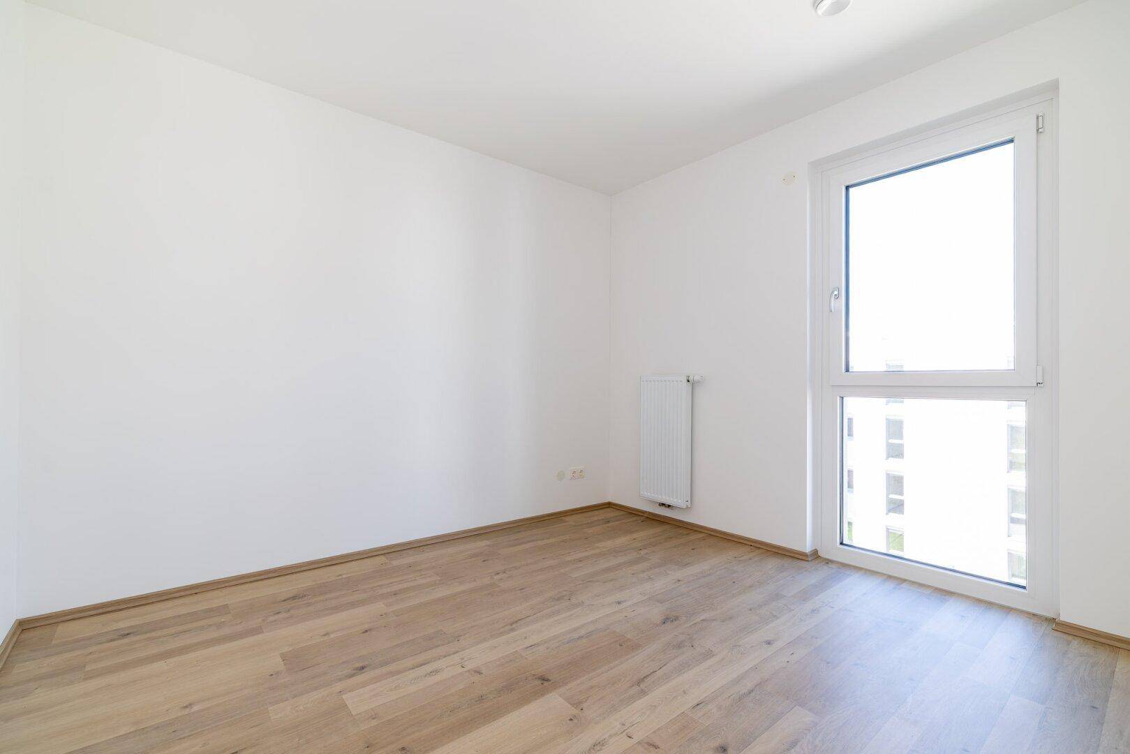 Heller Raum mit Laminatboden und einem hohen Fenster, das viel Tageslicht hereinlässt.