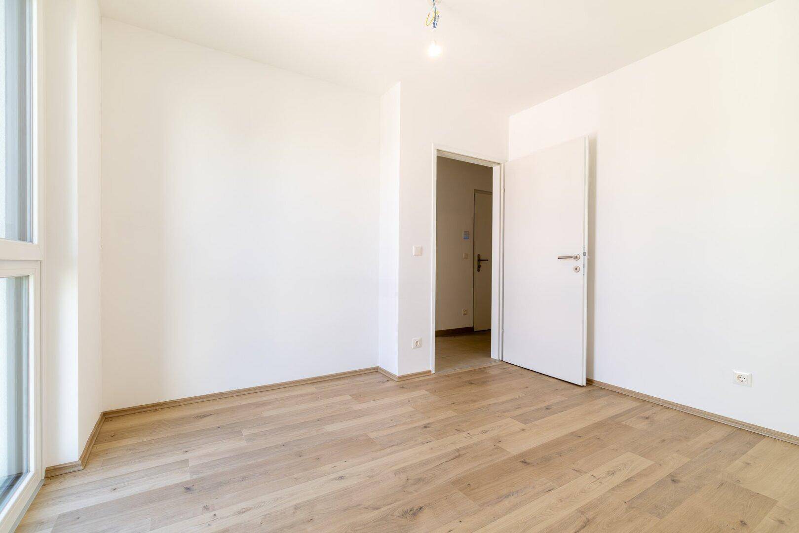 Leeres Schlafzimmer mit Laminatboden und einer weißen Tür, die zu einem weiteren Raum führt.