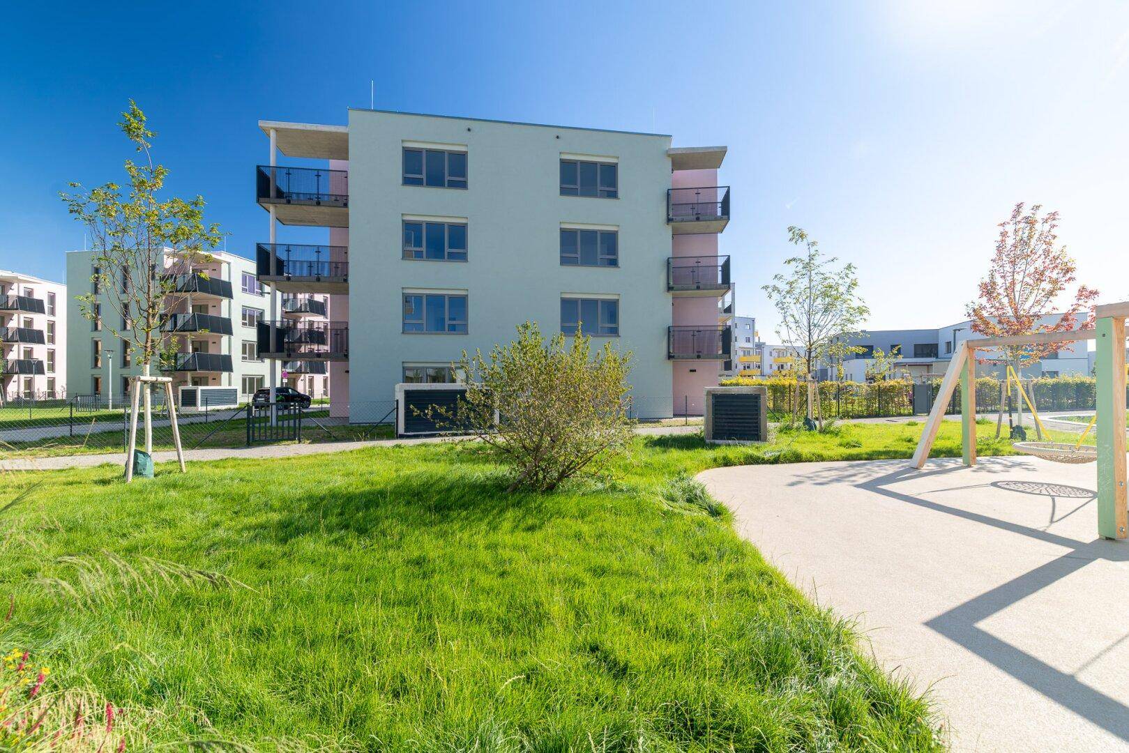 Moderne Wohnanlage mit Balkonen, Grünflächen und einem Spielplatz unter strahlend blauem Himmel.