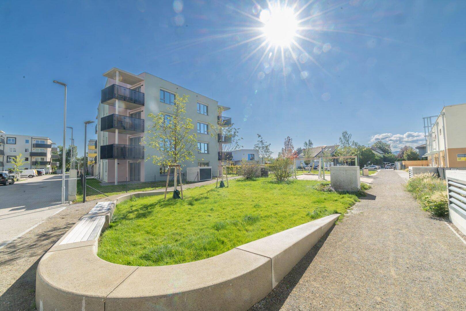 Sonnige Außenansicht eines modernen Wohnkomplexes mit Grünflächen und Sitzgelegenheiten.