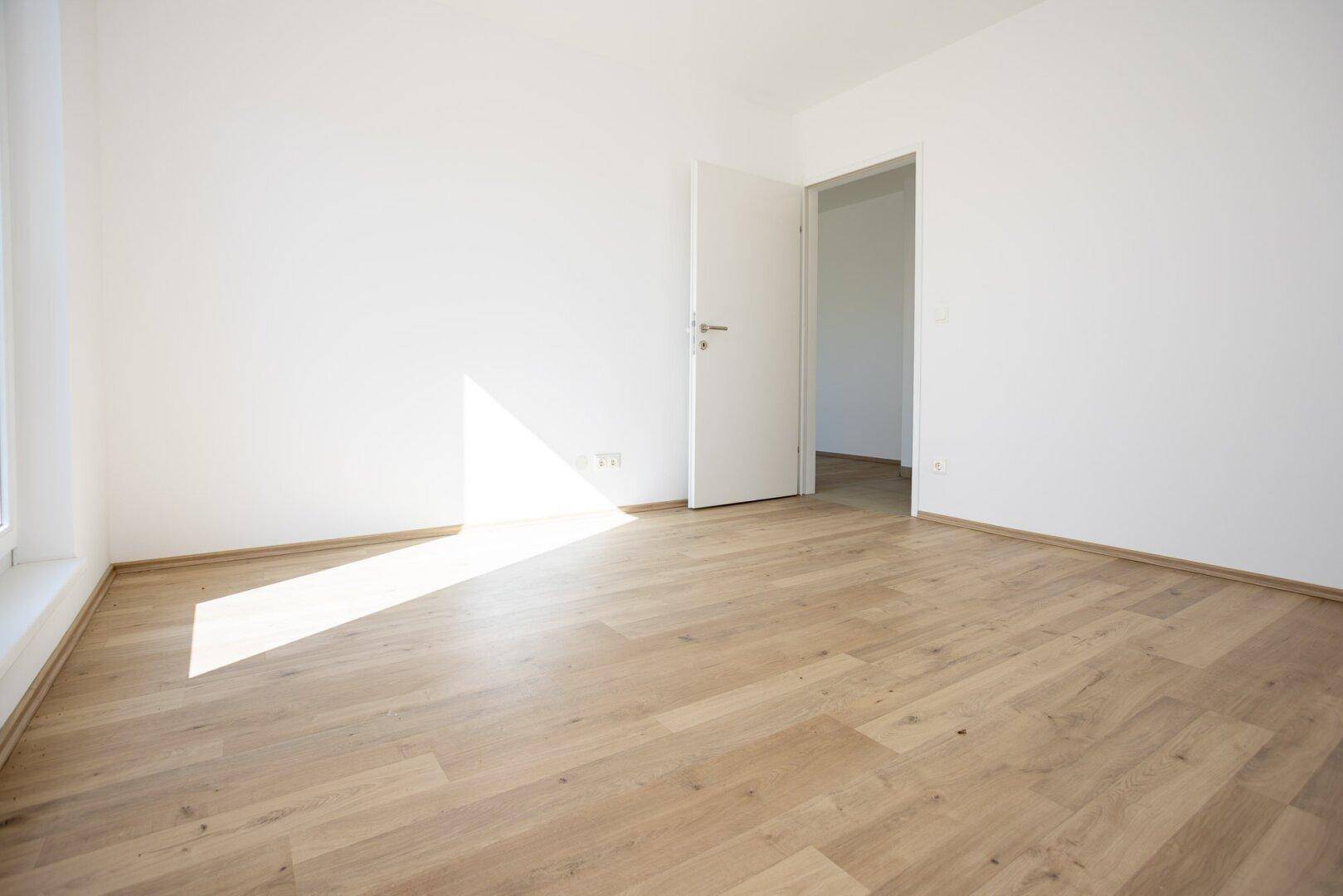 Geräumiges Zimmer mit Laminatboden, weiß gestrichenen Wänden und viel Sonnenlicht.