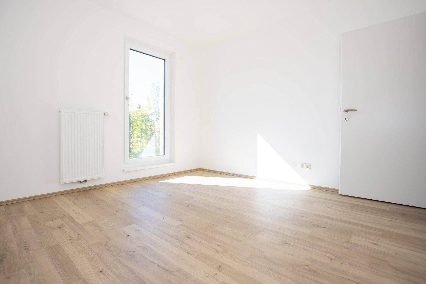Freundliches Zimmer mit Laminatboden, großem Fenster und Blick ins Grüne.