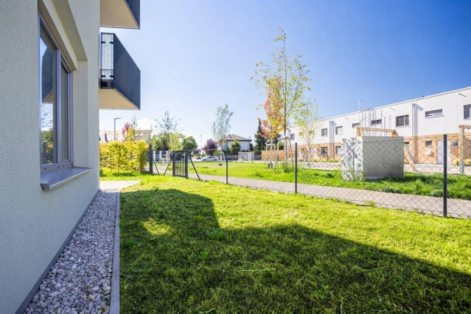 Weitläufige Rasenfläche neben dem Gebäude mit Kiesstreifen und Blick auf die Wohnanlage.
