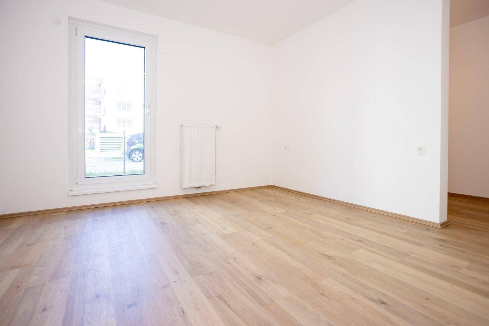 Helles, leeres Zimmer mit Laminatboden und einem hohen Fenster, bereit zur individuellen Gestaltung.
