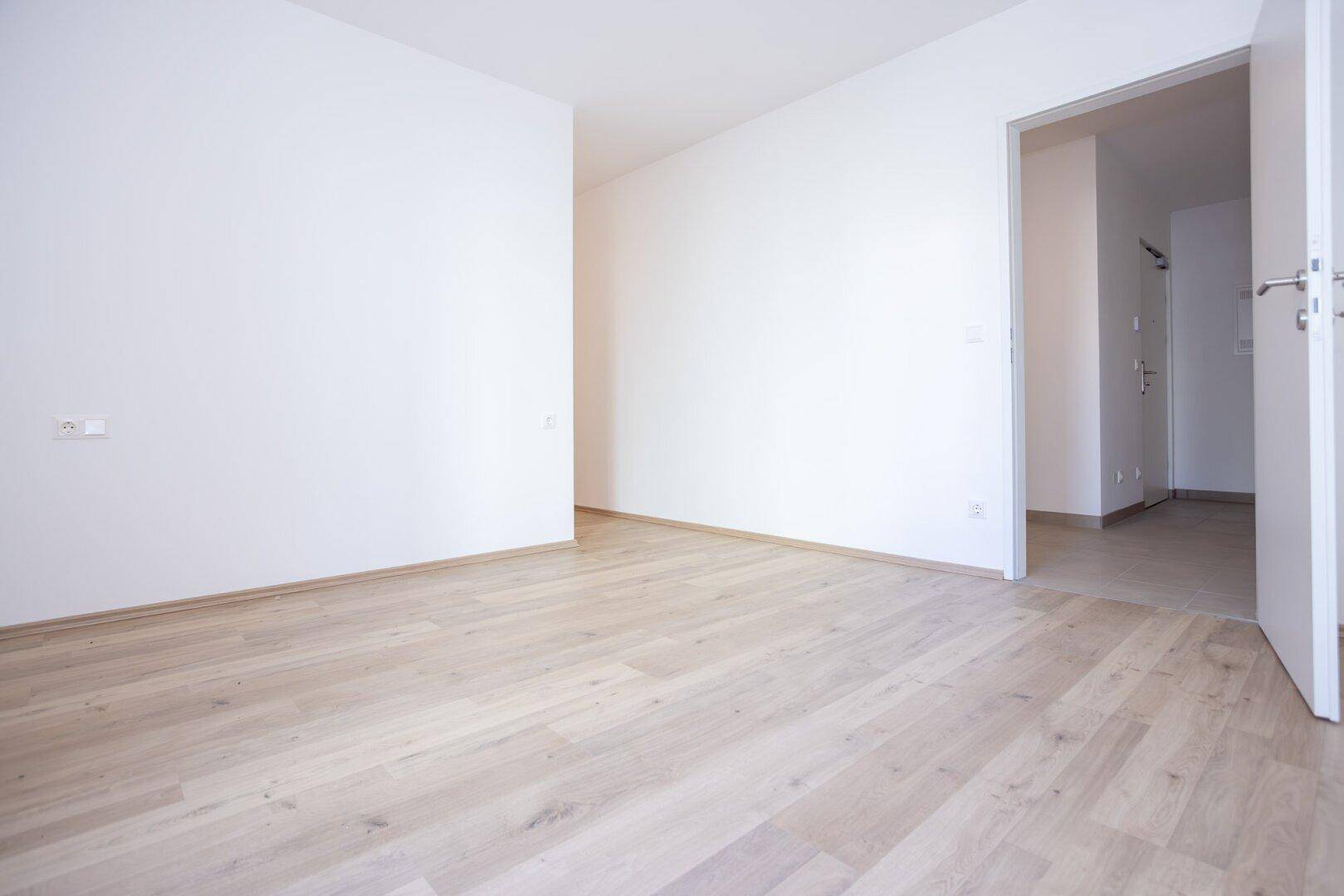 Leeres Zimmer mit hellem Laminatboden und Zugang zu einem weiteren Raum oder Flur.