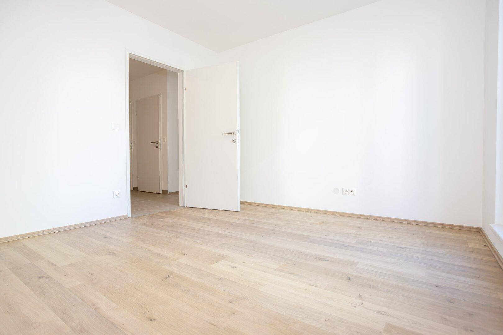 Helles, leeres Zimmer mit Laminatboden und offener Tür zu einem angrenzenden Bereich.
