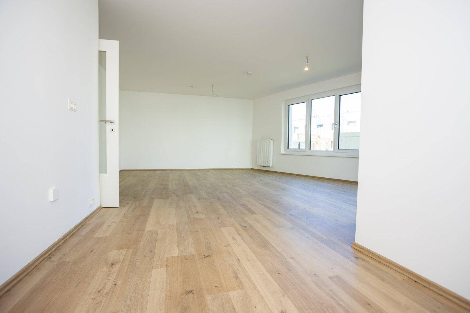 Leeres Wohnzimmer mit hellem Laminatboden und einem großen Fenster, das viel Licht hereinlässt.
