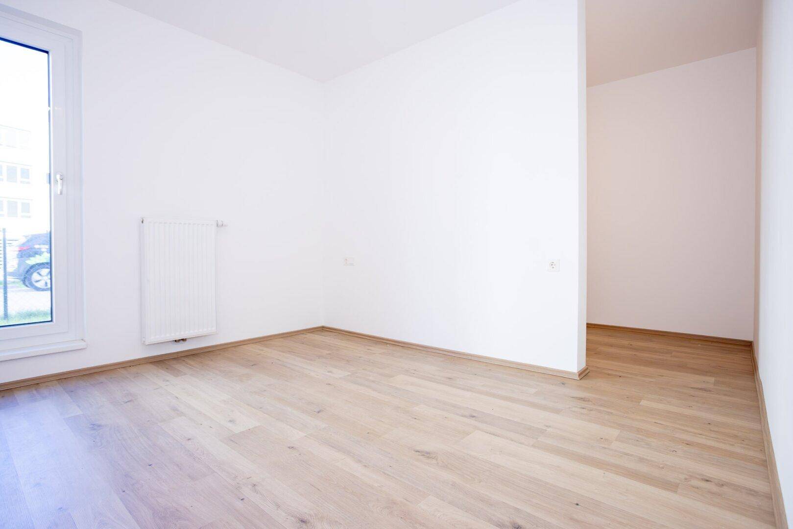 Leeres Zimmer mit hellem Laminatboden und einem Fenster, das Tageslicht spendet.