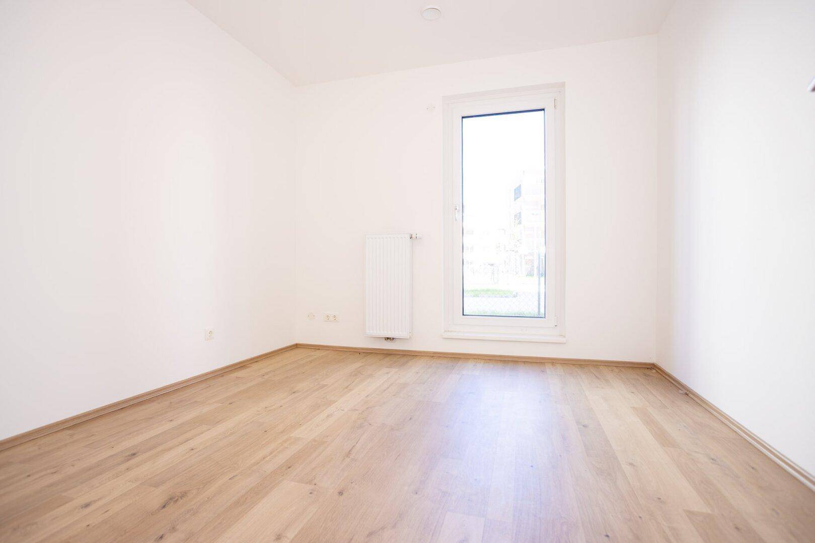 Helles, leeres Zimmer mit Laminatboden und einem hohen Fenster, ideal für ein Schlafzimmer.