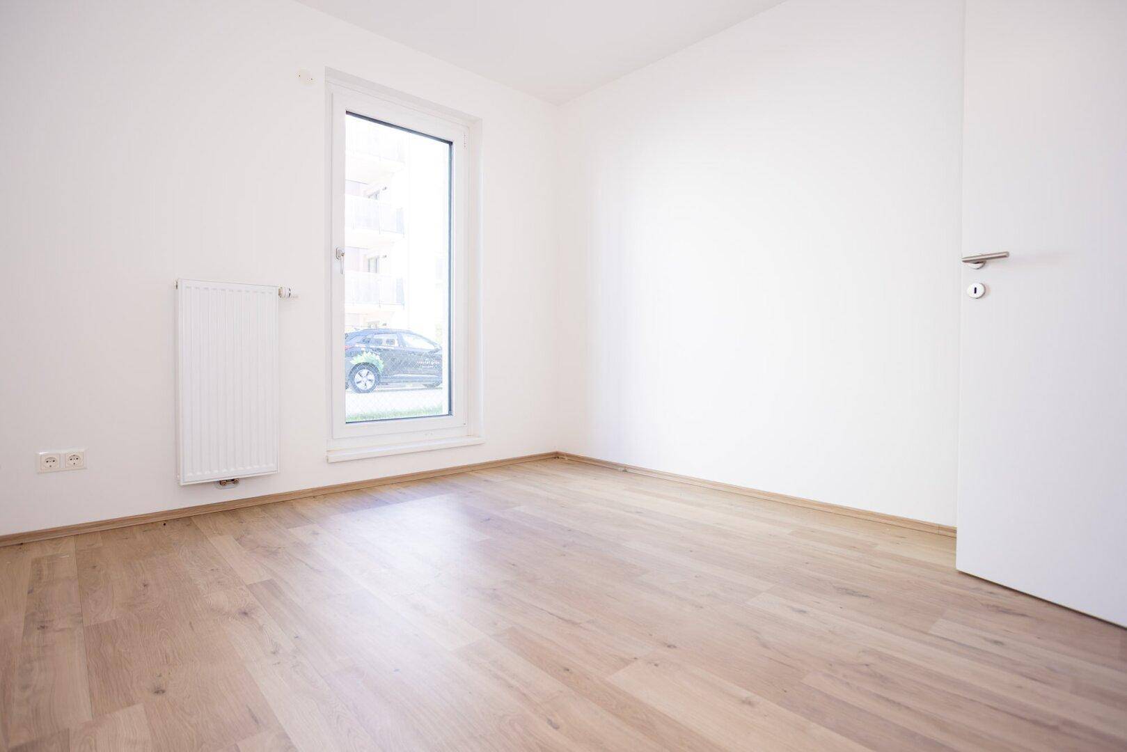 Leeres Zimmer mit hellem Laminatboden und einem hohen Fenster, das viel Licht hereinlässt.