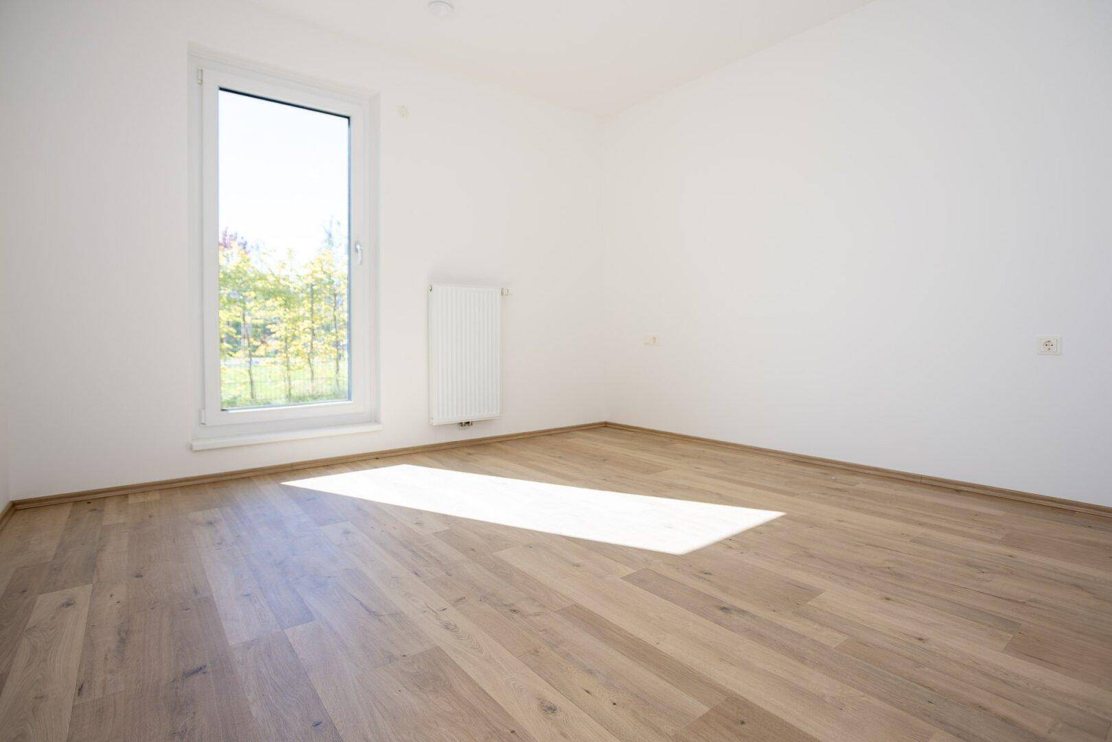 Leeres Schlafzimmer mit hellem Laminatboden und großem Fenster, das viel Licht hereinlässt.