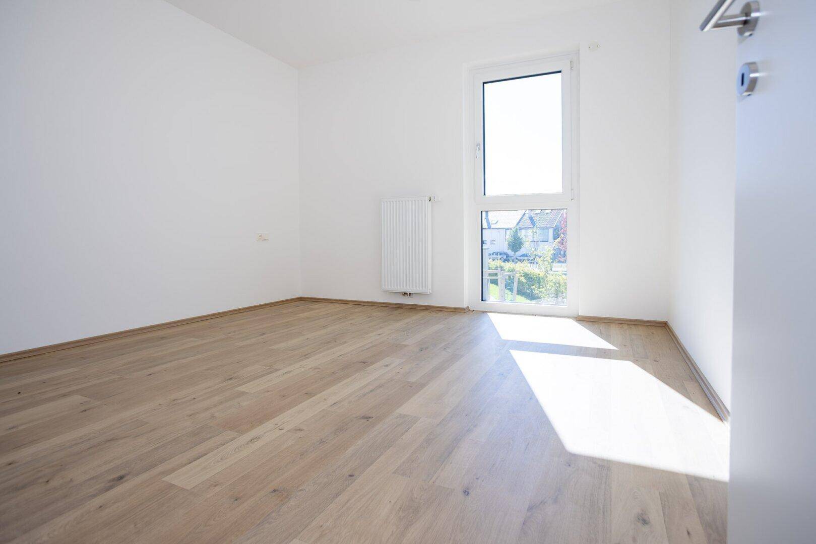 Helles Schlafzimmer mit Laminatboden, großem Fenster und Heizkörper, unmöbliert.