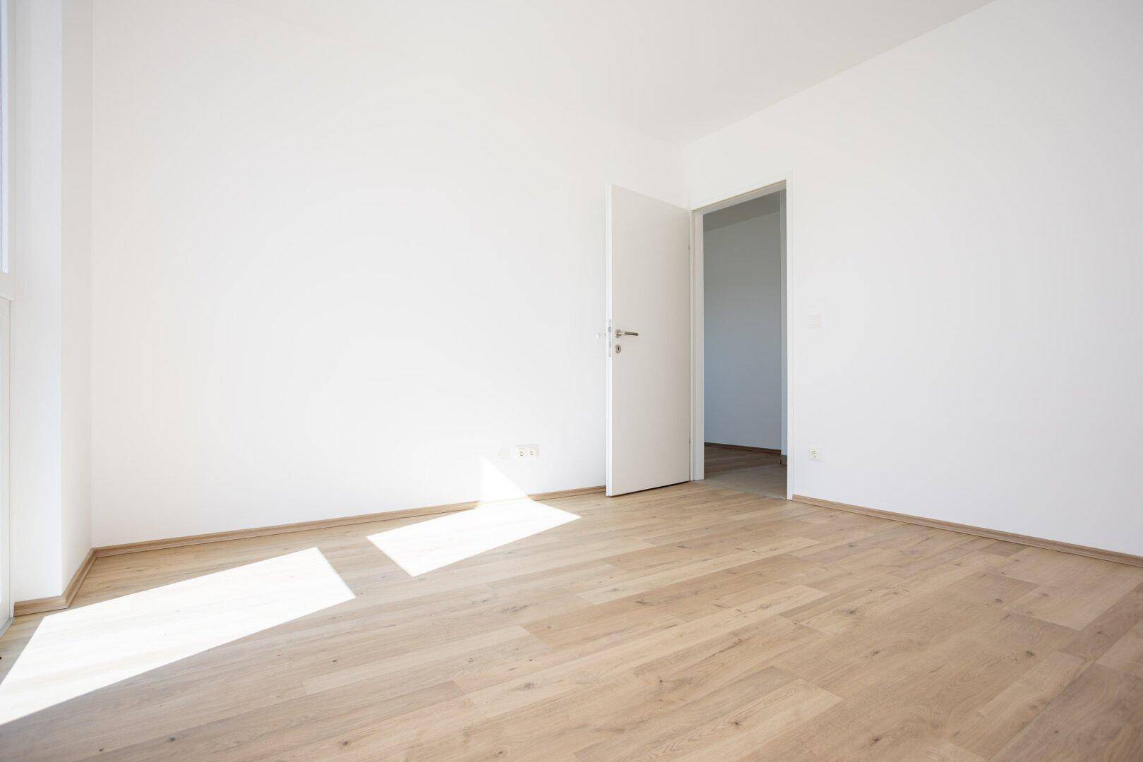Unmöbliertes Schlafzimmer mit Laminatboden, weißer Tür und viel natürlichem Licht.